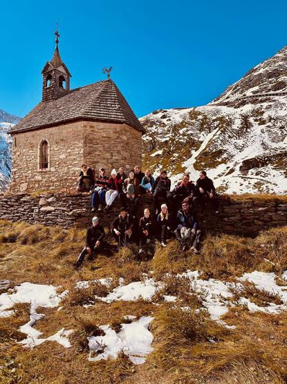The excursion group in front of an old stone chapel at the Tauernfenster. There are patches of snow on the grass and surrounding mountain peaks.