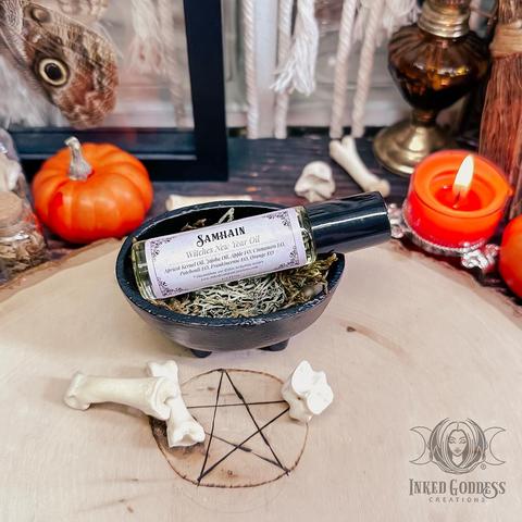 A small round vial of yellowish Samhain Witches New Year Oil is propped in a small black cauldron atop a bed of herbs. It's surrounded by animal bones, tiny pumpkins, and a tealight candle. It has a black cap and a label that lists its ingredients. This is an exclusive product from Inked Goddess Creations.