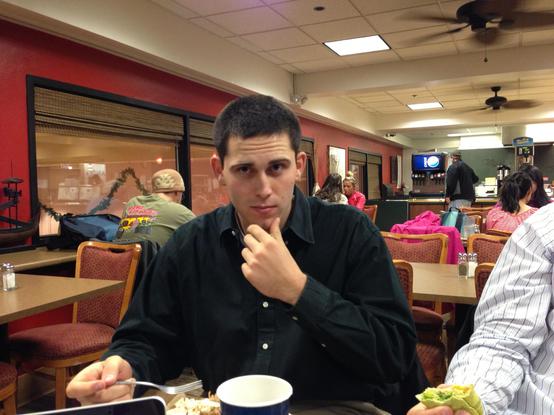 A young man with short hair (Matt Boyd) is seated at a table in a restaurant, thoughtfully resting his chin on his hand. He is wearing a black shirt and has a plate of food in front of him. In the background, other diners are visible, with