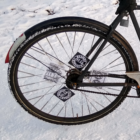 Man sieht das Hinterrad eines schwarzen Fahrrads auf schneebedeckter Straße. Es sind Spike-Reifen aufgezogen und in den Speichen stecken schwarze und weisse Speichenkarten mit dem Logo der RespectCyclists.