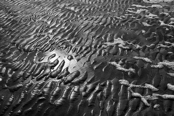 Eine S/W-Aufnahme: Die Brandung der Nordsee hat flächige wulstige Erhebungen in den Sandstrand geformt. Auf einigen Erhebungen befindet sich noch weißer Schaum, die kleinen Pfützen zwischen den Erhebungen reflektieren das Sonnenlicht.