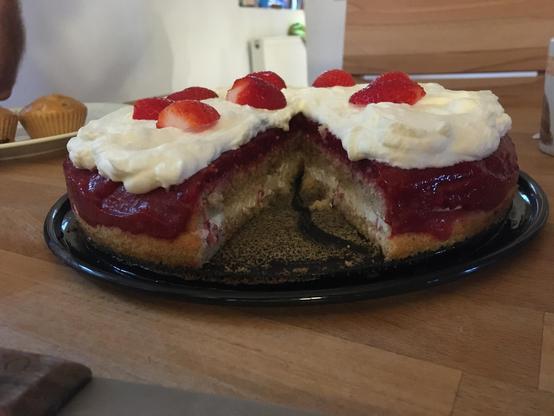 sponge cake visible from the side: halved cake filled with a layer of plain yoghurt, topped with bright red mashed strawberries, whipped cream and decorated with bright red strawberry halves