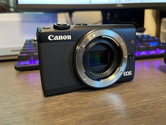 A black Canon EOS M100 camera with no lens mounted. The camera’s APS-C sensor is visible at the center of the lens mounted. 