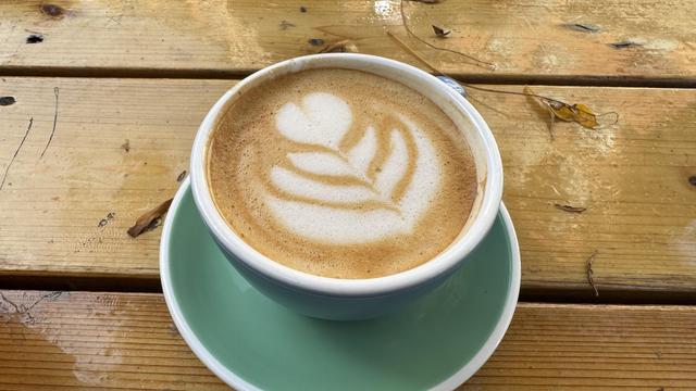 Photo of a latte drink topped with latte art in a white and teal cup on top of a saucer, resting on a wooden table made of several boards with gaps in between them. The table appears wet reflecting a rainy day.