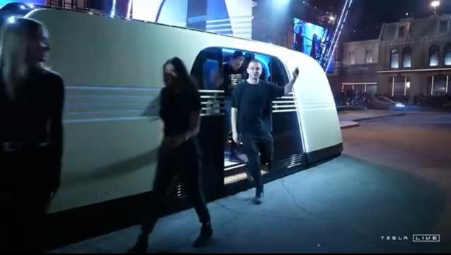 people wave as they step down out of a futuristic looking gold vehicle parked by a kerb in an urban setting at night