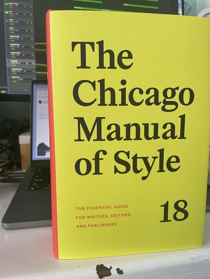a book standing on a desk in front of a laptop and monitor. Cover is bright yellow, title is The Chicago Manual of Style 18.