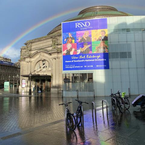 Usher Hall with a rainbow overhead, some bikes, colorful reflections on wet pavement 
