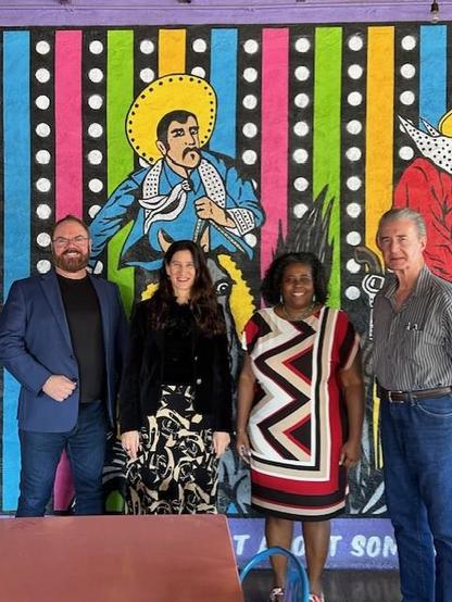 Drs. Dean Colston, Annette Padilla, Jane Garcia, and Raymond Hopkins are posing for the camera at a Mexican restaurant. The background is a wall painting of a festive and colorful image that includes a man in a sombrero. 