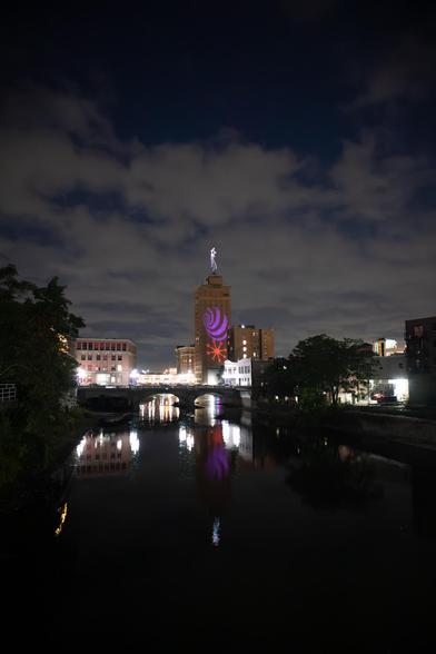 A night scene of downtown Aurora, Illinois. The Fox River is in the foreground, with a well-lit bridge crossing it in the center of the image. A tall building (Leland Tower) with some colorful lights projected on it stands behind the bridge.