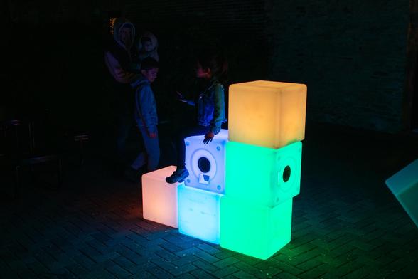 Some kids playing on brightly lit blocks arranged in a staircase formation. One child is sitting on one of the steps while the other is standing on the ground looking at her.