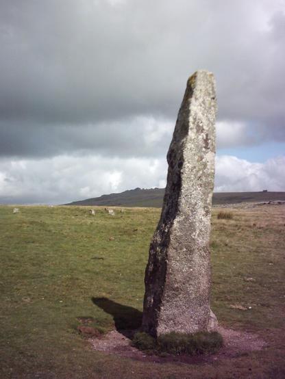 The 3.1m or just over 10ft tall menhir at Merrivale near Princetown on Dartmoor, with a slight lean. A stone circle lies just over 40m to the north and is between about 18 and 20.5m diameter. The grass on the moor is short and sheep-clipped with the tors in the distance below a grey cloudy sky.