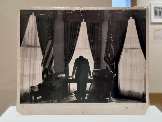 JFK leaned over the Oval Office desk on his hands, facing away. Three windows light the image and put him in in silhouette.
