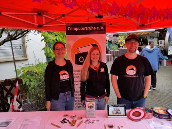 Foto eines Infostandes mit Merch, Werbematerial, einem Infodisplay und Keksdosen. Dahinter stehen zwei Frauen und und ein Mann, lächelnd in Vereinsklamotten.