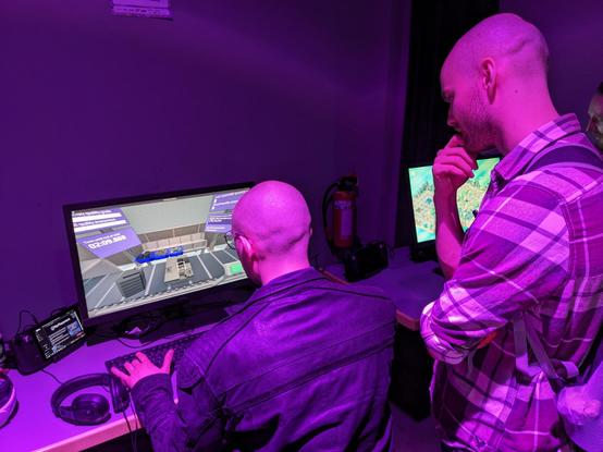 Person sitting at desk with Steam Deck, a monitor, mouse and keyboard, playing the Re:Placement demo, with another person watching over their shoulder