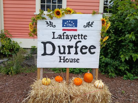 Charming hand painted sign in front of the Lafayette Durfee house. Some sunflowers are vined around the top and side. A hay bale with 5 fall gourds sits beneath it.