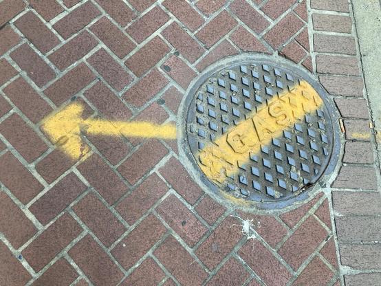Manhole cover with a diamond design and the inscription „Gas“, the middle part is spray-painted yellow. A yellow arrow is sprayed across the surrounding sidewalk.