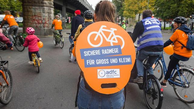 Eine Person fährt auf einer Familien-Fahrraddemo mit dem Fahrrad. Auf ihrem Rücken ist ein Demoschild angebracht, das im Zentrum des Bildes steht: "ADFC: Sichere Radwege für Groß und Klein!"