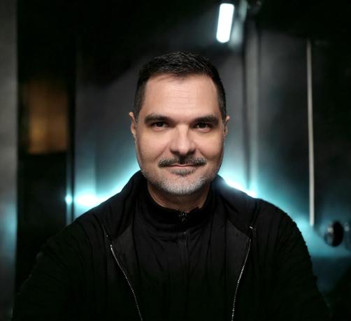 Porträt des DJs Niko Ruhm aka Grille. Er trägt eine schwarze Jacke und schaut etwas verschmitzt direkt in die Kamera. Der zeigt Hintergrund unscharf metallische Wände, die bläulich-grün beleuchtet sind.