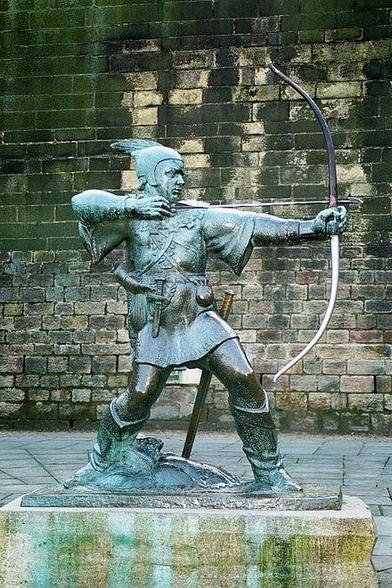 A bronze statue of a warrior archer aiming with a bow, set against a textured stone wall. The figure is depicted in a dynamic pose, wearing traditional attire with a quiver at the side.  It's Robin Hood. In Nottingham. 