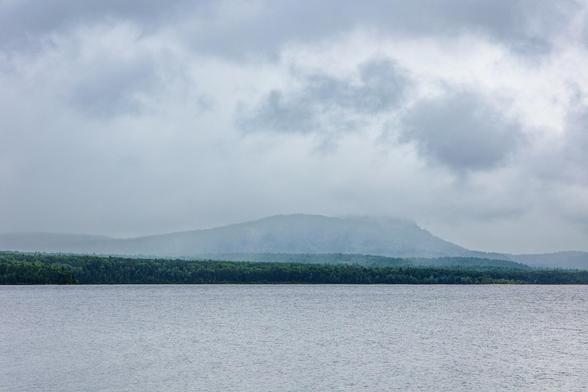 A calm river is framed by a distant tree line and a misty mountain under a cloudy sky.