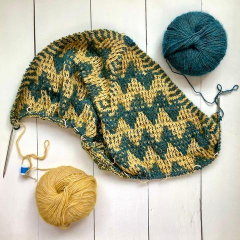 Flat lay view of a triangular shawl with mosaic patterning being knit on a circular needle. It’s shown with a ball of yellow and a ball of petrol colored yarn on a white plank background.