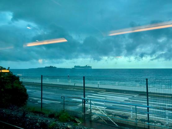 A coastal scene featuring a dark, cloudy sky over the ocean. Two ships are visible in the distance along with a coastal fence and road in the foreground.