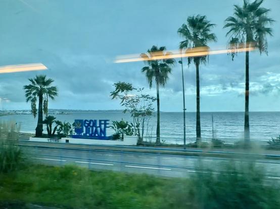 A coastal view featuring palm trees and a sign for Golfe Juan, with the sea visible in the background under a cloudy sky. The image is taken from a train whose orange neon lights are reflected in the window and superimposed on the photo. 