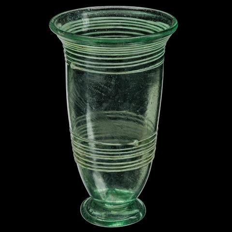 Hoher schmaler grüner Glasbecher mit Standfuss, verziert mit umlaufenden Wülsten unter dem ausschwingenden Rand und im unteren Drittel. Fotografiert vor schwarzem Hintergrund.