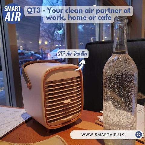  A compact QT3 air purifier on a café table next to a glass water bottle. Text: ‘Your clean air partner for home, work, or café.’