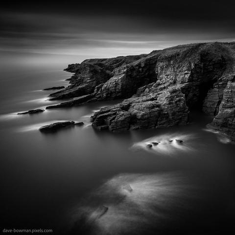 A black and white photograph titled "Arbroath Cliffs" features the rugged, textured cliffs of Arbroath in Angus, Scotland. The long exposure effect softens the surrounding elements, giving the sea a smooth, misty appearance while the cliffs remain sharp and prominent. The image focuses on the natural contours and dramatic lines of the cliffs, highlighting their raw beauty against the tranquil atmosphere created by the monochrome tones.