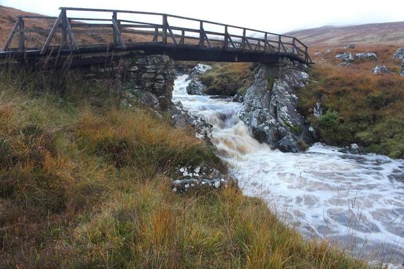 A wooden bridge with stone supports crossing a stream which is in space, long exposure to blur the water. The ground either side is grassy with some rocks.