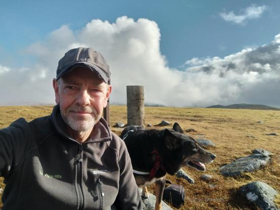 Selfie, me and Motray the dog. We are sat next to a concrete pillar, the summit trig point. The hilltop is mostly grassy, there is a stony cairn next to the trig point. Blue sky and clouds.