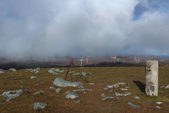 View from the summit. There is a concrete pillar trig point, and a bit of iron that looks like a broken fence post. The hill top is grassy with some boulders. In ther middle distance are about 20 wind turbines. Cloudy, with the mist down to the ground in the distance.