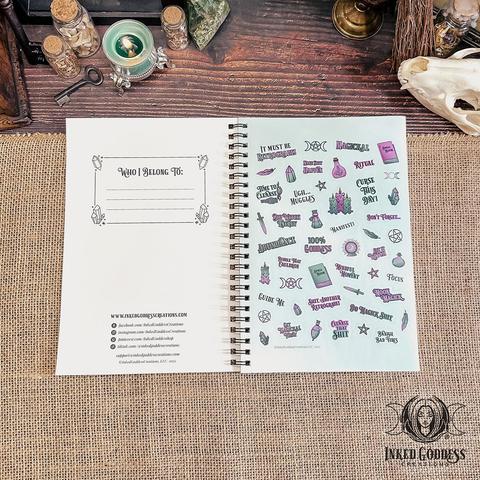 An open spiral-bound journal lies on a burlap-covered surface. The inside cover features a bookplate and copyright information. The facing page is a sheet of purple and teal stickers with witchy vibes to use in the "Busy Witch 2025 Monthly Date Planner." This is an exclusive product from Inked Goddess Creations.