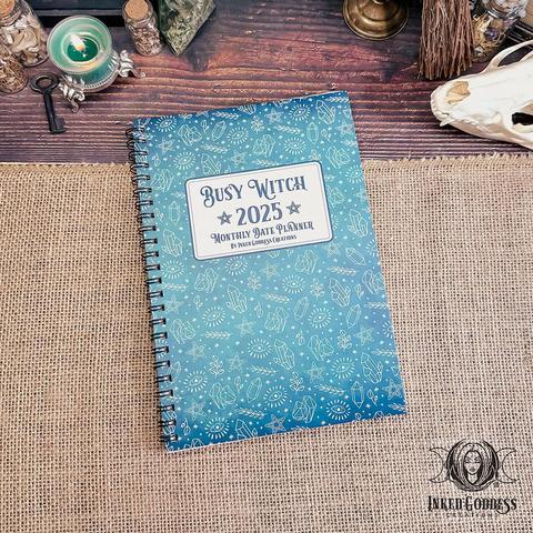 A blue spiral-bound journal lies closed on a burlap-covered surface. The background image of the cover is a gradient of blues and there's a white block in the center with the title "Busy Witch 2025 Monthly Date Planner." This is an exclusive product from Inked Goddess Creations.