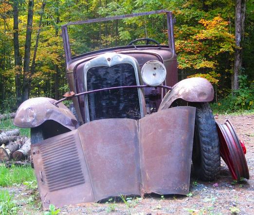 A rusty old truck sits in the woods with colorful fall foliage in the background.