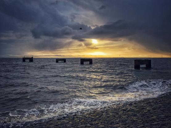 Vier dunkle Betonpfeiler stehen hintereinander in der etwas unruhigen Nordsee. Der Himmel ist stark bewölkt, lässt aber bei tiefstehender Sonne etwas golden erscheinendes Licht durch, das sich auch im Wasser widerspiegelt. 