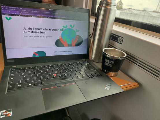 Aufgeklappter Laptop mit der Startseite von my-friday.org ("Ja, du kannst etwas gegen die Klimakrise tun."), daneben eine Thermoskanne und ein Becher, rechts Blick aus dem Zugfenster. 