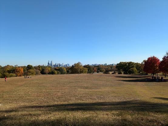 Photo: A broad expanse of grassy field with a row of multihued trees behind it and the skyline of downtown and west Philadelphia behind that. There are some people on the lawn. The sky is bright blue.