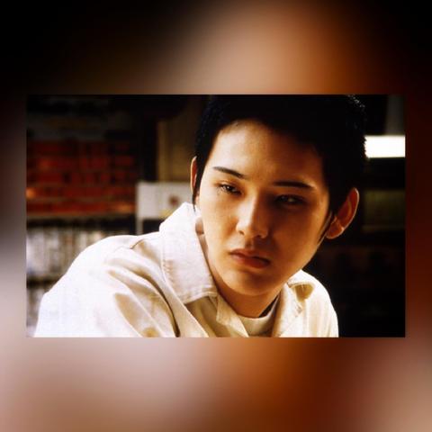 Film still from the movie 9 SOULS in a colored box frame. 

The image is of a young Japanese man wearing a white collared shirt with a white t-shirt beneath it. He is looking down and to his right, across his body, and wears a blank, impassive look on his face.