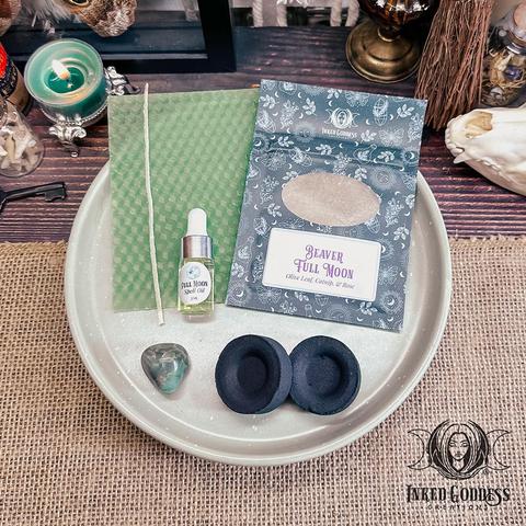 The contents of our exclusive "Beaver Full Moon Ritual Kit for the November 15th, 2024, Full Moon in Taurus" product are arranged on a small tray. They include a sheet of green beeswax and a wick; a pouch of Beaver Full Moon herbs, including olive leaf, catnip, and rose; a dropper vial of our exclusive Full Moon Spell Oil; a tumbled emerald gemstone; and two charcoal discs. Presented by Inked Goddess Creations.