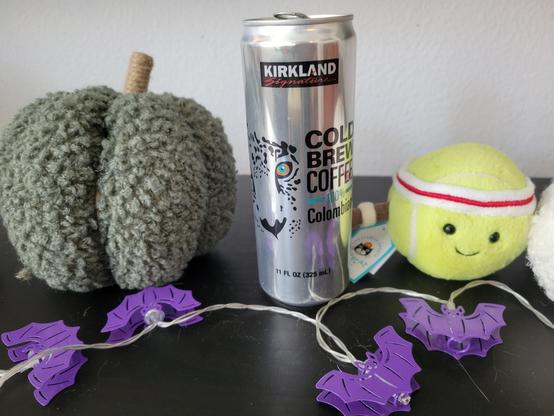 Can of Kirkland Signature Colombian Cold Brew Coffee, 11 fl oz (6-inches tall, 2-inches diameter). The can is shiny aluminum. It has the Kirkland Signature logo at top (black rectangular background, "Kirkland" in white block letters, "Signature" in red script). An illustration of the face of a leopard is at left-center, next to text "Cold Brew Coffee 100% Colombian" in black block letters (100% is in green-blue). Footer text is "11 FL OZ (325 mL)" in black font.

Several other objects are arranged on the surface of an IKEA HEMNES TV unit (black-brown stain). At left is a green Boucle Fabric Pumpkin (World Market). At right is an Amuseables Sports Tennis Ball plush (Jellycat). In the foreground is an unlit string of purple-colored bat lights.