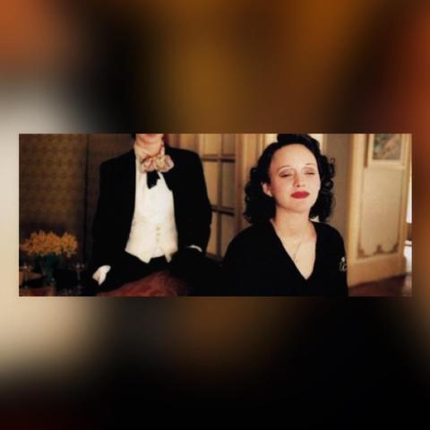 Film still from the movie LA VIE EN ROSE in a colored box frame. 

The image is of a woman with dark, curly, shoulder-length hair and a black dress looks forward with her eyes half closed. A female character in male B&W outfit stands behind her, to the left, with hands in pockets facing her back.