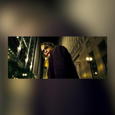 Film still from the movie THE DARK KNIGHT in a colored box frame.

The image is a low-angle shot of the character of the Joker as played by Heath Ledger. He has mussed, greasy green hair, sloppy clown makeup with a wide mouth, and wears a dark purple. His face is turned to the left in the direction of the camera, otherwise he is in profile. Two tall, Gotham City buildings loom in the background on both sides of him. He splits the image.