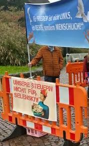 Zu sehen ist eine Kundgebung mit einer Detailansicht. An einer orangefarbenen Baustellenabseprrung ist ein Plakat angebracht. Es zeigt die Märchenfigur Pinoccio, die eine Schale voller Geldscheine hält. Beschriftet ist das Plakat mit der in Versalien aufgebrachten Botschaft "Unsere Steuern nicht für Selensky!. Hinter der Absperrung steht ein Mann, dessen Gesicht durch ein weiteres Plakat verdeckt ist. Auf dem blauen Banner steht: "Für FRIEDEN ist nicht LINKS, nicht RECHTS, sondern MENSCHLICH!" Eine weiße Taube flattert auf den Schriftzug zu. 
