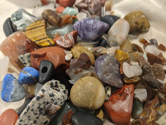 A mixed pile of slightly shiny, multicolored rocks on a off white tea towel. Most of the rocks are shades of brown, with a few blues, purples and reds. Also spots and stripes. I don't know the names yet.