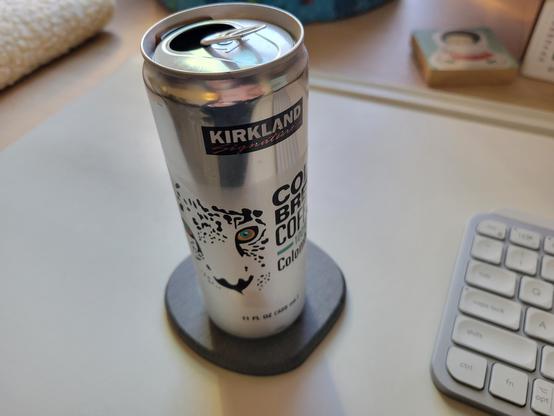 Can of Kirkland Signature Colombian Cold Brew Coffee, 11 fl oz (6-inches tall, 2-inches diameter), on a gray diatomaceous-earth coaster on an off-white desk mat on a maple desk. The can is shiny aluminum. It has the Kirkland Signature logo at top (black rectangular background, "Kirkland" in white block letters, "Signature" in red script). An illustration of the face of a leopard is at left-center, next to text "Cold Brew Coffee 100% Colombian" in black block letters (100% is in green-blue). Footer text is "11 FL OZ (325 mL)" in black font.