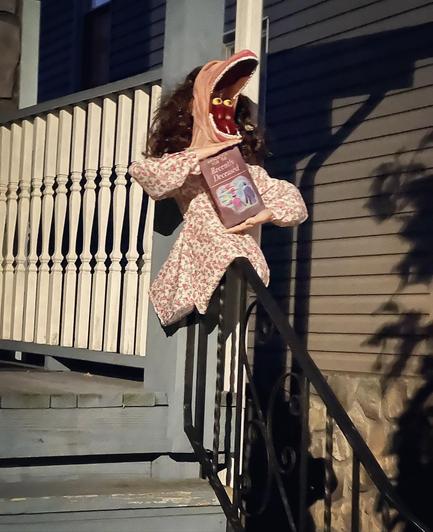 Barbara from Beetlejuice sitting on a porch bannister holding the Handbook for the Recently Deceased. Her head is distorted into the wide mouth and eyeballs sitting on the back of her tongue pose. 