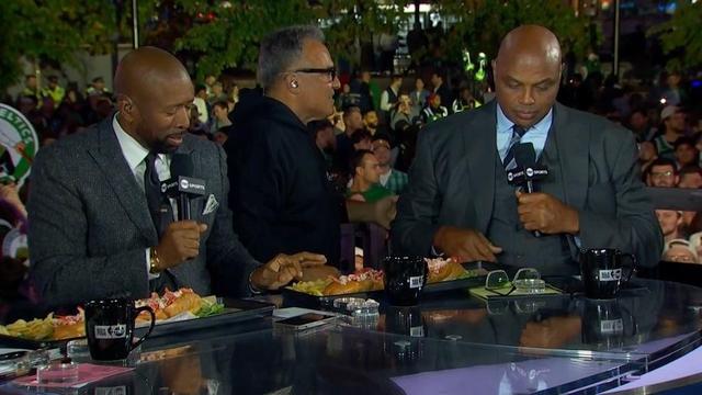 Two men are seated at a table with food in front of them, discussing on a live broadcast. One man is holding a microphone while the other is looking down at a notepad. The background features a crowd of people and greenery, indicating an outdoor