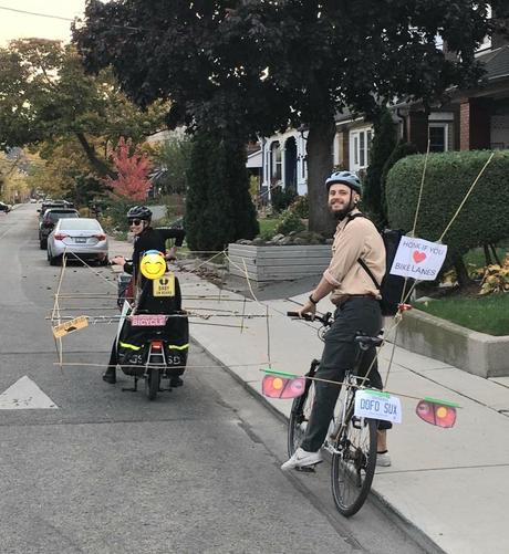 Two people riding bikes with bamboo poles to make the bikes look bigger and like cars. They have bumper stickers that say "baby on board", "I brake for turtles", "my other car is also a bicycle", "honk if you ❤️ bike lanes" and a fake licence plate that says "DOFO SUX"